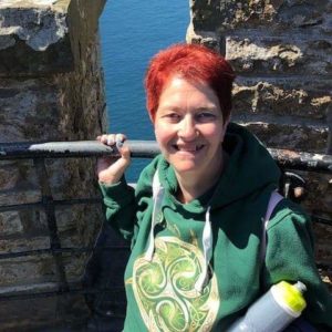 Cynthia Owens is a white woman with short-cropped red hair. She's leaning against the rail between two stone pillars that show a hint of pretty blue water.