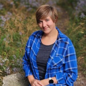 Leah Lore is a woman with short-cropped brown hair. She is sitting on a large boulder in a field of wildflowers.